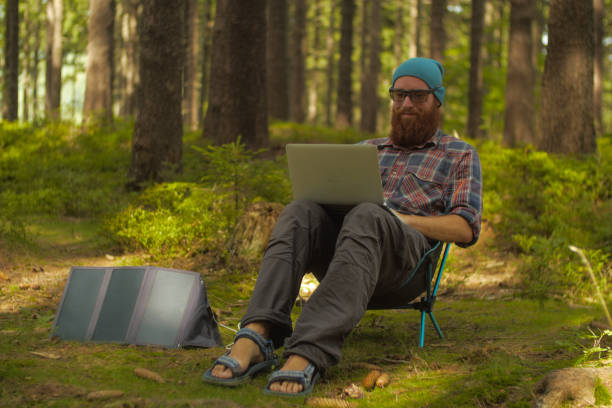 A man camping in the woods charging his laptop with a solar panel