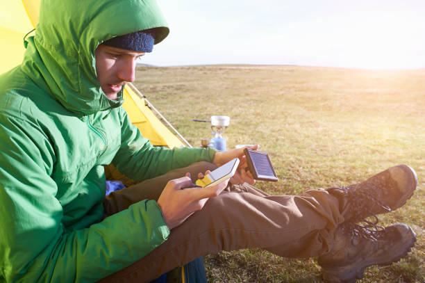 A man camping in the wilderness on a moorland sits in a tent and charges his smartphone using a portable solar panel.